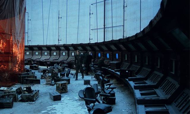 A large abandoned room with old computer terminals lining the circular perimeter. The floor is covered in debris and partially-disassembled computers. Three people dressed in black investigate the scene.